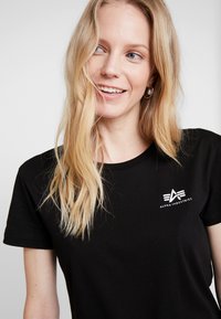 Schwarzes Baumwoll-T-Shirt mit Rundhalsausschnitt und kurzen Ärmeln, mit einem weißen Logo im linken Brustbereich. Glatte Textur, taillierte Passform.