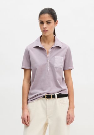 Vrouw met donker haar in een staart, gekleed in een lichtpaarse polo met korte mouwen, ingestopt in een witte broek met een zwarte riem.