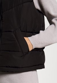 Černá péřová vesta s prošívaným designem a přední kapsou, nošená přes světle šedý žebrovaný svetr, s hladkým, texturovaným povrchem.