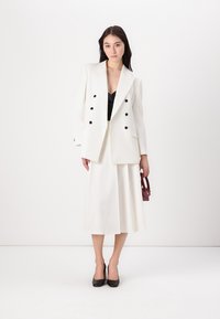 White tailored blazer with black buttons paired with a white skirt and black heels. A small burgundy handbag is held in one hand.