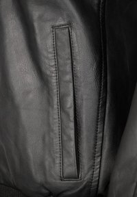 Primo piano di una giacca di pelle nera, che mostra la superficie liscia, le cuciture e un dettaglio della tasca orizzontale sul lato sinistro.