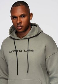 Nuori mies, jolla on lyhyet hiukset, pukeutuneena vaaleanharmaaseen huppariin, jossa on mustat nauhat ja rinnassa teksti "ombre wear".