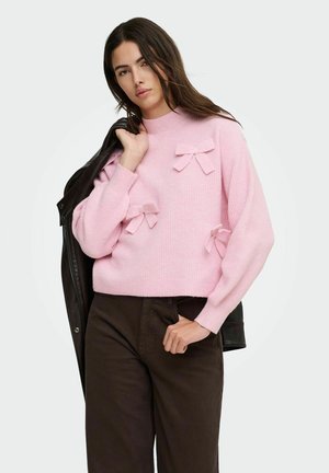 Femme portant un pull rose avec trois nœuds en tissu, tenant une veste noire sur l'épaule et portant un pantalon foncé, sur un fond uni.