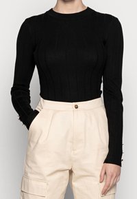 Top negro de manga larga con tejido acanalado, cuello redondo y silueta ajustada; presenta botones decorativos en los puños de las mangas. Pantalones beige de talle alto.