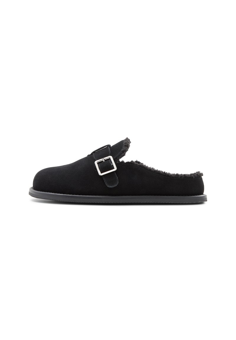 Svart mocka slip-on tofflor med ett spänne, foder av shearling och gummisula; rund tå och utan baksida.