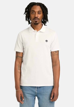 Uomo con dreadlocks che indossa una polo bianca a maniche corte con piccolo logo sul petto e jeans azzurri, in piedi di fronte a uno sfondo semplice.