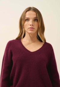 Burgundowy sweter z dekoltem w serek, o prążkowanej teksturze i długich rękawach. Miękka dzianina, luźny krój, z wyraźnym przeszyciem wzdłuż dekoltu.