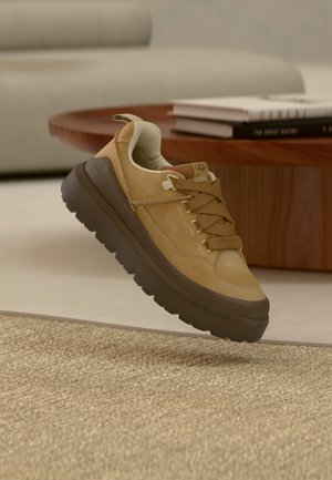 Beżowy zamszowy sneaker z brązową gumową podeszwą, wyposażony w teksturowane sznurowadła i wyściełany kołnierz. Podniesiony design z subtelnym detalem marki.