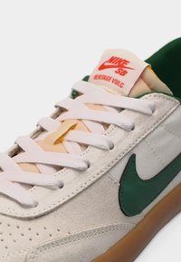 Κοντινή λήψη μπεζ παπουτσιού Nike SB Heritage Vulc με λευκά κορδόνια, σκούρο πράσινο λογότυπο Nike και σόλα από καουτσούκ.
