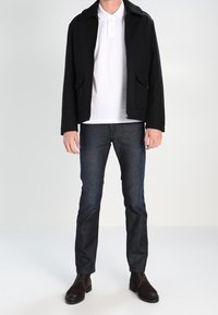 Veste noire avec un col en fourrure, polo blanc en dessous, jeans foncés, et chaussures à lacets marron. Design simple et coupe ajustée.