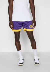 Shorts de baschet purpurii cu accente galbene, din material tip plasă, cu logo pe piciorul stâng, purtați cu șosete albe și tenisi alb-negru.