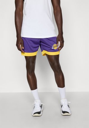 Pantaloni basketball viola con accenti gialli, tessuto a rete, logo sulla gamba sinistra, indossati con calzini bianchi e sneakers in bianco e nero.