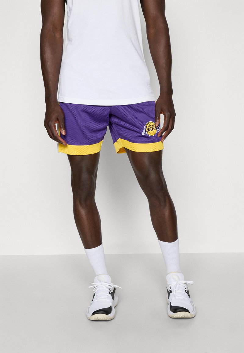 Shorts de baschet purpurii cu accente galbene, din material tip plasă, cu logo pe piciorul stâng, purtați cu șosete albe și tenisi alb-negru.