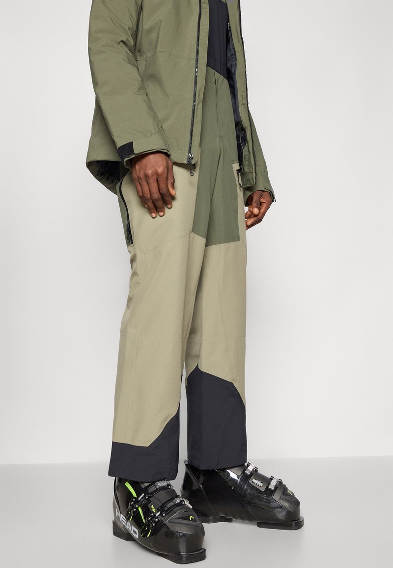 Spodnie narciarskie w oliwkowo-zielonym i beżowym kolorze, z zamkiem kolorowym i teksturowymi panelami, w połączeniu z czarnymi butami narciarskimi.