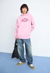 Personne debout sur un sol bleu, portant un sweat à capuche New Balance rose, un jean bleu ample, des baskets vertes, tenant un sac fourre-tout noir à son côté.