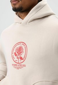 Beige hoodie gemaakt van zacht materiaal, met een rode rozenafbeelding en de tekst "Leef Authentiek, Hou Passioneel" en de merknaam "Young Atelier."