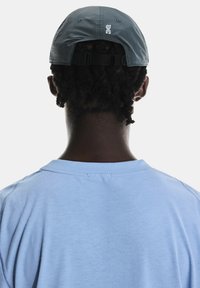 Căciulă gri cu curea ajustabilă, purtată pe dos. Tricou bleu deschis cu o textură moale, cu linii de cusătură vizibile și un croi lejer la guler.
