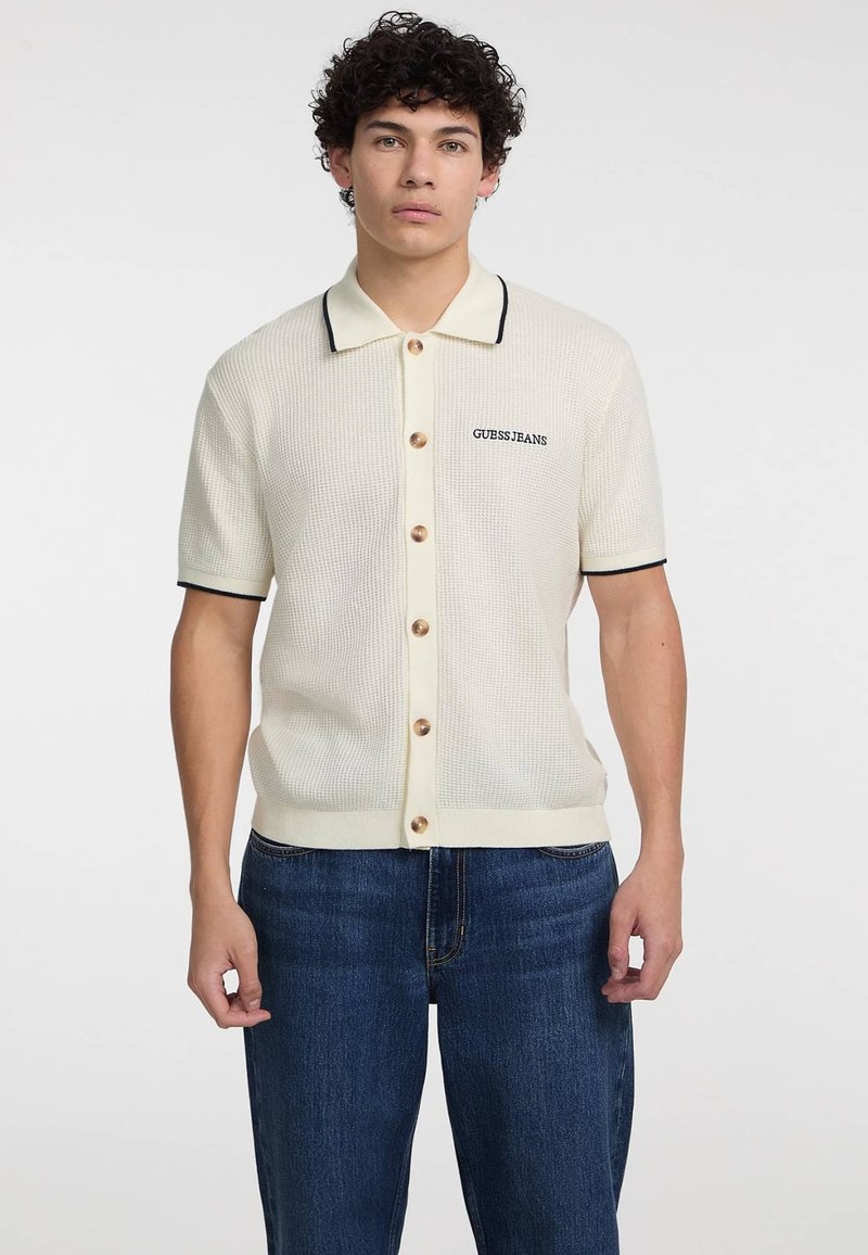 Cremfarbener gerippter Poloshirt mit braunen Knöpfen, schwarzem Besatz am Kragen und den Ärmeln, mit "GUESS JEANS"-Logo auf der linken Seite.