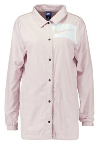 Světle růžová bunda Nike s límcem, zapínáním na druky, elastickými manžetami, dvěma bočními kapsami a překryvným logem na levé straně hrudi.