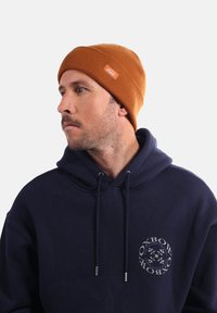 Bonnet tricoté orange avec une texture côtelée et un petit patch logo. Porté par une personne en sweat à capuche bleu marine avec un détail de logo circulaire.