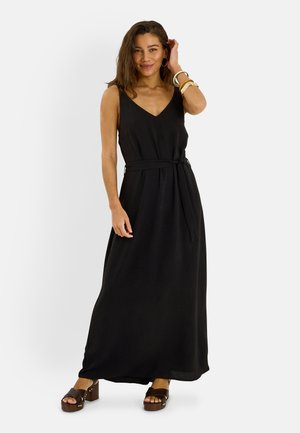 Mujer con un vestido negro largo sin mangas con un cinturón atado a la cintura y sandalias marrones de plataforma, sonriendo y tocándose el cabello.