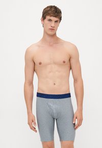 Giovane uomo con capelli castani corti che indossa boxer lunghi grigi con una fascia blu, in piedi davanti a uno sfondo bianco semplice.