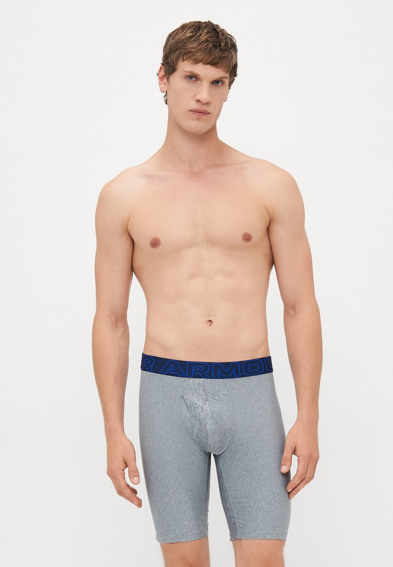 Giovane uomo con capelli castani corti che indossa boxer lunghi grigi con una fascia blu, in piedi davanti a uno sfondo bianco semplice.