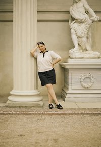 Kratki rukavi, svijetložuti pleteni top s crnim ovratnikom, u paru s crnom suknjom. Stojeći pored klasičnog bijelog stupa i statue.