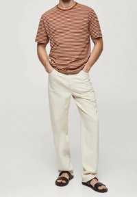 Camisa de manga corta a rayas en marrón y crema, pantalones de algodón blancos y sandalias negras. La camisa tiene un escote redondo y un corte holgado.