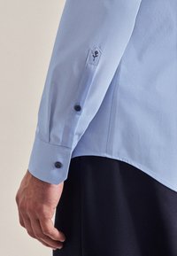 Camicia azzurro chiaro a maniche lunghe con polsini abbottonati e ricamo di un fiore sul braccio. Presenta bottoni scuri e una texture liscia.