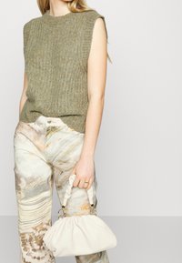 Mouwloze groene ribgebreide top, gecombineerd met beige en crèmekleurige wijd pijpbroek met een patroon; houdt een witte leren pouch tas met gouden accenten vast.