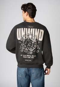 Schwarzes Sweatshirt mit lockerer Passform, auf der Rückseite mit einem großen cremefarbenen Grafikdesign. Der Text beinhaltet "UNWIND" sowie Kritzeleien von Wolken und Händen.