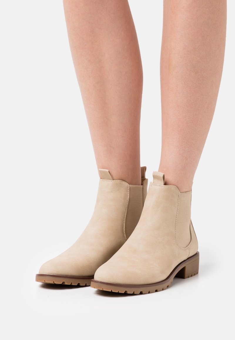 Bottines en daim beige clair avec talons bas et carrés et panneaux latéraux élastiques portées sur des jambes nues, présentées sur un fond blanc.