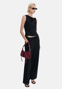 Top nero a maglia con design asimmetrico e dettaglio annodato, abbinato a pantaloni neri larghi. Gli accessori includono una piccola borsa bordeaux e sandali neri.