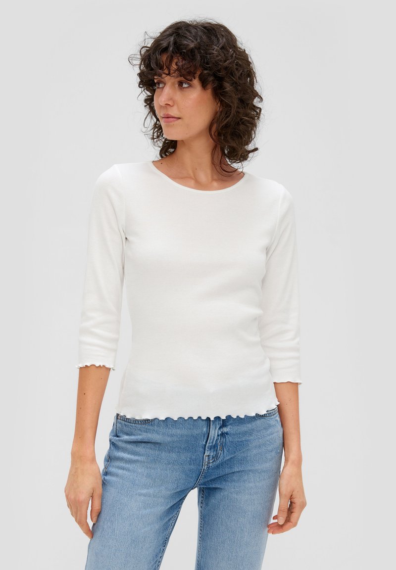 Witte geribbelde lange mouw top met een ronde hals en een scalloped zoom. Heeft driekwartmouwen en een aansluitend ontwerp. Gecombineerd met lichtblauwe jeans.