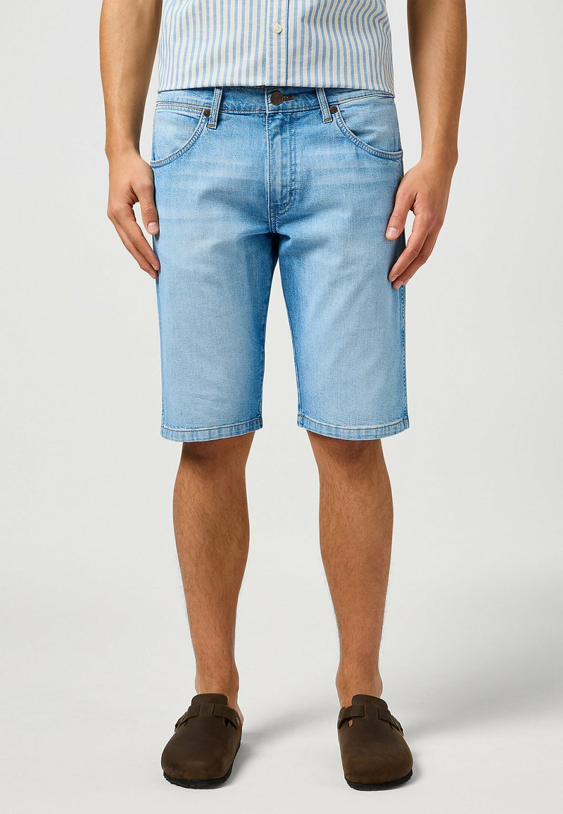 Pantalones cortos de mezclilla azul claro con corte recto, cinco bolsillos y dobladillos enrollados. Combinados con zapatos marrones de tipo slip-on y una camiseta a rayas.