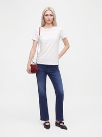 Femme aux longs cheveux blonds portant un t-shirt blanc, un jean bleu, des ballerines noires pointues, et tenant un sac rouge perlé avec une bandoulière en chaîne.