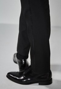 Zapatos de vestir negros con puntera redondeada y diseño de cordones, combinados con pantalones negros ajustados que presentan un detalle en la costura lateral. Textura suave, ajuste moderno.