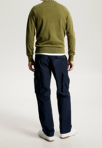 Pull vert olive avec un col rond et des poignets côtelés, associé à un pantalon cargo marine avec plusieurs poches et une jambe fuselée. Baskets blanches.