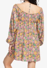 Femme portant une robe florale colorée à manches longues, tatouage vertical dans le dos, et collier de coquillages sur fond blanc.