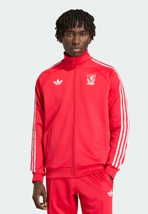 Mężczyzna ubrany w czerwony dres Adidas z herbem Liverpool Football Club, stojący z jedną ręką w kieszeni na jednolitym tle.