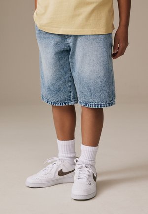 Barn iført lyseblå denimshorts, hvide ankelsokker, hvide Nike-sneakers med brun swoosh og en gul skjorte, stående på en neutral baggrund.