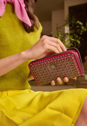 Hand die een portemonnee met patroon en ritssluiting vasthoudt, draagt een gele rok en een groen topje, zittend binnen met planten op de achtergrond.