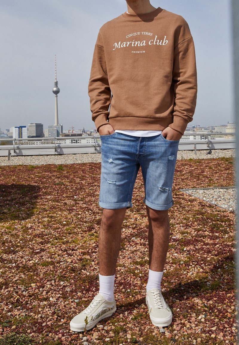 Sudadera marrón con texto blanco, pantalones cortos de mezclilla azules con dobladillos enrollados y zapatillas beige con acentos decorativos, con un fondo de ciudad.