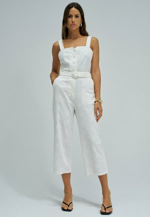 Femme portant une combinaison blanche sans manches ceinturée avec une texture florale et des boutons, debout les mains dans les poches devant un fond uni.
