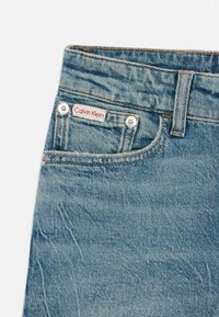 Svetlo modré džíny z denimu s klasickým dizajnom piatich vreciek, kontrastným prešívaním a červenou logom nášivkou na páse.