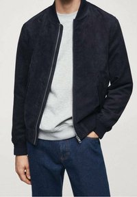 Giubbotto bomber in camoscio blu navy con cerniera frontale, polsini e orlo a costine, abbinato a una felpa grigio chiaro e jeans di denim blu.