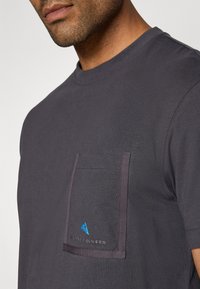 Tricou din bumbac gri închis cu decolteu rotund, dotat cu un buzunar pe piept cu o bordură contrastingă și un accent de logo albastru. Textură netedă.