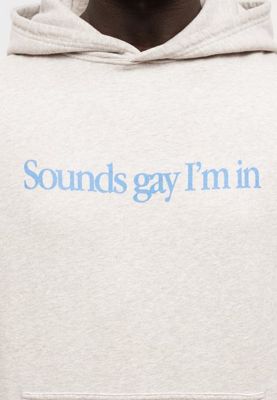 Svijetlosiva majica s kapuljačom s prednjim džepom, s plavim natpisom "Sounds gay I'm in." Materijal izgleda mekan s glatkom teksturom.
