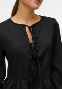 Juodas suknelė su tekstūruotu audiniu, turinti V formos iškirptę ir surišimo detalę priekyje. Ilgos rankovės ir susiraukšlėta sijono dalis.
